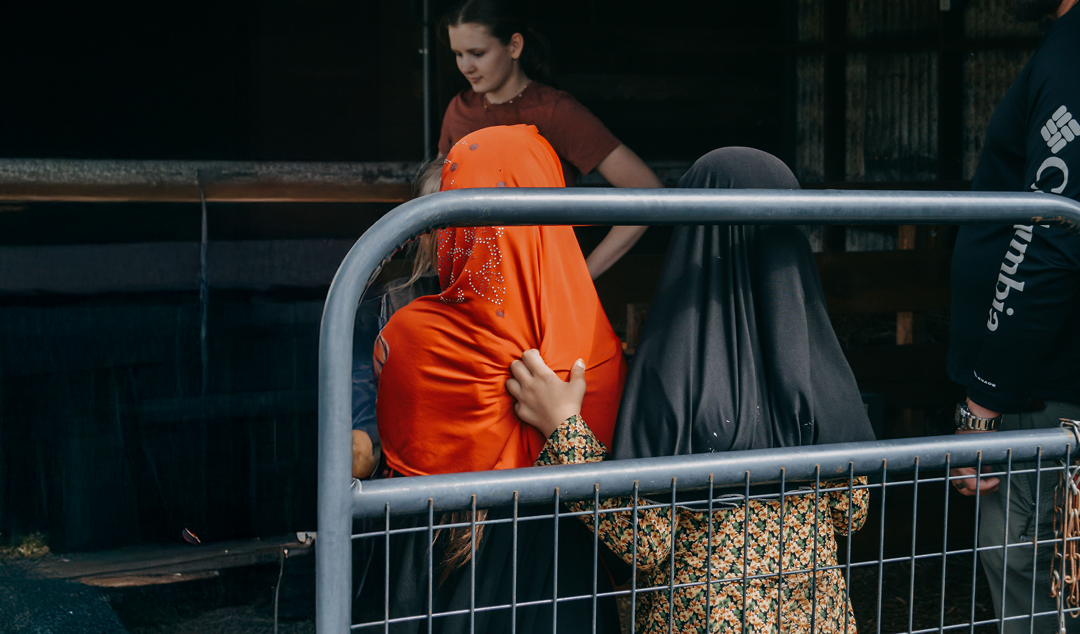 Three people near a metal fence, two in headscarves, one in long-sleeved black shirt. A fourth person is blurred in the background.