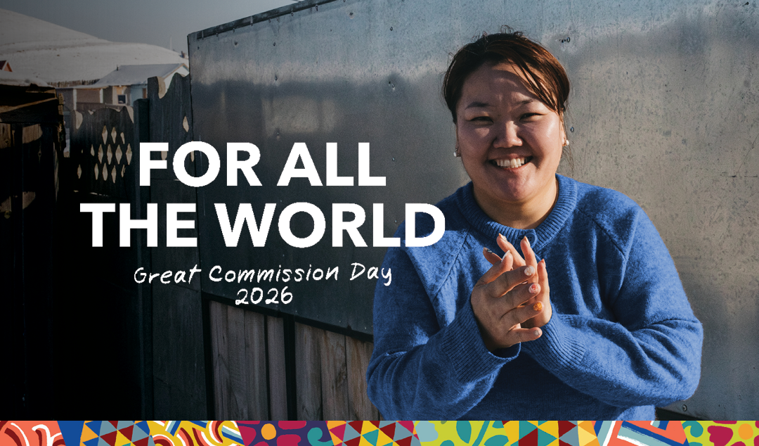 Text reading ‘For All the World – Great Commission Day 2026’ over an outdoor background with a person in a blue sweater and a colorful patterned border at the bottom.