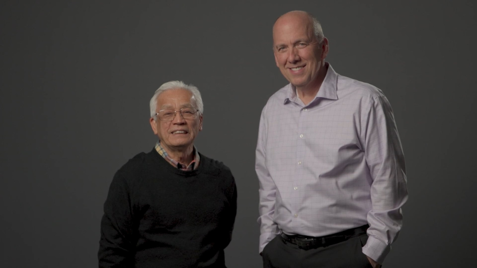 Two individuals standing side by side against a plain dark background. The person on the left is wearing a dark sweater, and the person on the right is wearing a light-colored dress shirt with hands in pockets. The individuals are Vietnamese District Superintendent Linh Huynh and U.S. C&MA President Jonathan Schaeffer.