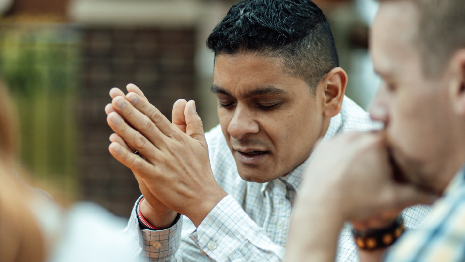 man sitting at a table praying with his hands together and eyes closed