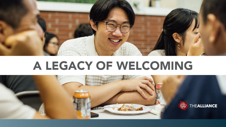 People sitting at a table with plates of food and canned drinks, with a banner across the center reading “A LEGACY OF WELCOMING” and The Alliance logo in the bottom right corner.