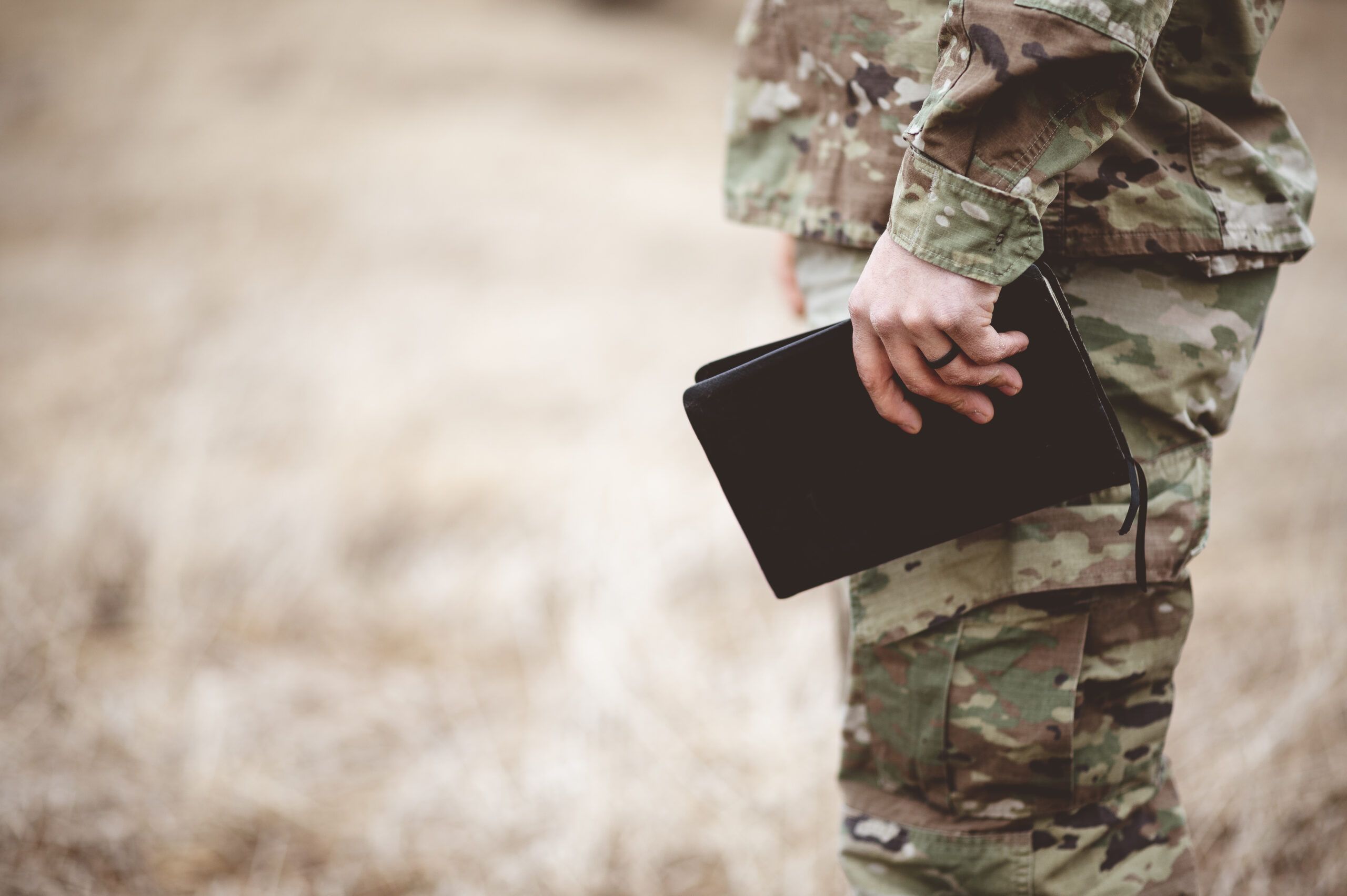 Close-up of a person in camouflage uniform holding a small black Bible while standing outdoors in a field.