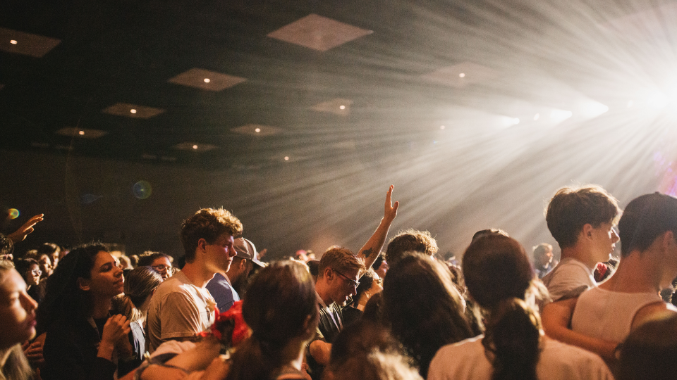 A large group of youth gathered closely together in a dimly lit indoor space, with beams of light streaming down from above. One person has an arm raised, and others are standing shoulder to shoulder, creating a sense of unity and engagement in worship. The background shows a ceiling with recessed lights and a warm, glowing atmosphere.