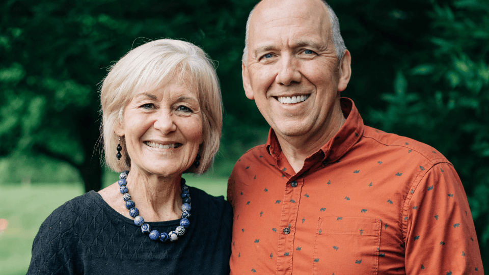 Jonathan and Mary Schaeffer standing outdoors in front of green foliage. Jonathan is wearing a rust-colored button-up shirt with a subtle pattern, and Mary is wearing a textured dark top with a chunky blue beaded necklace.