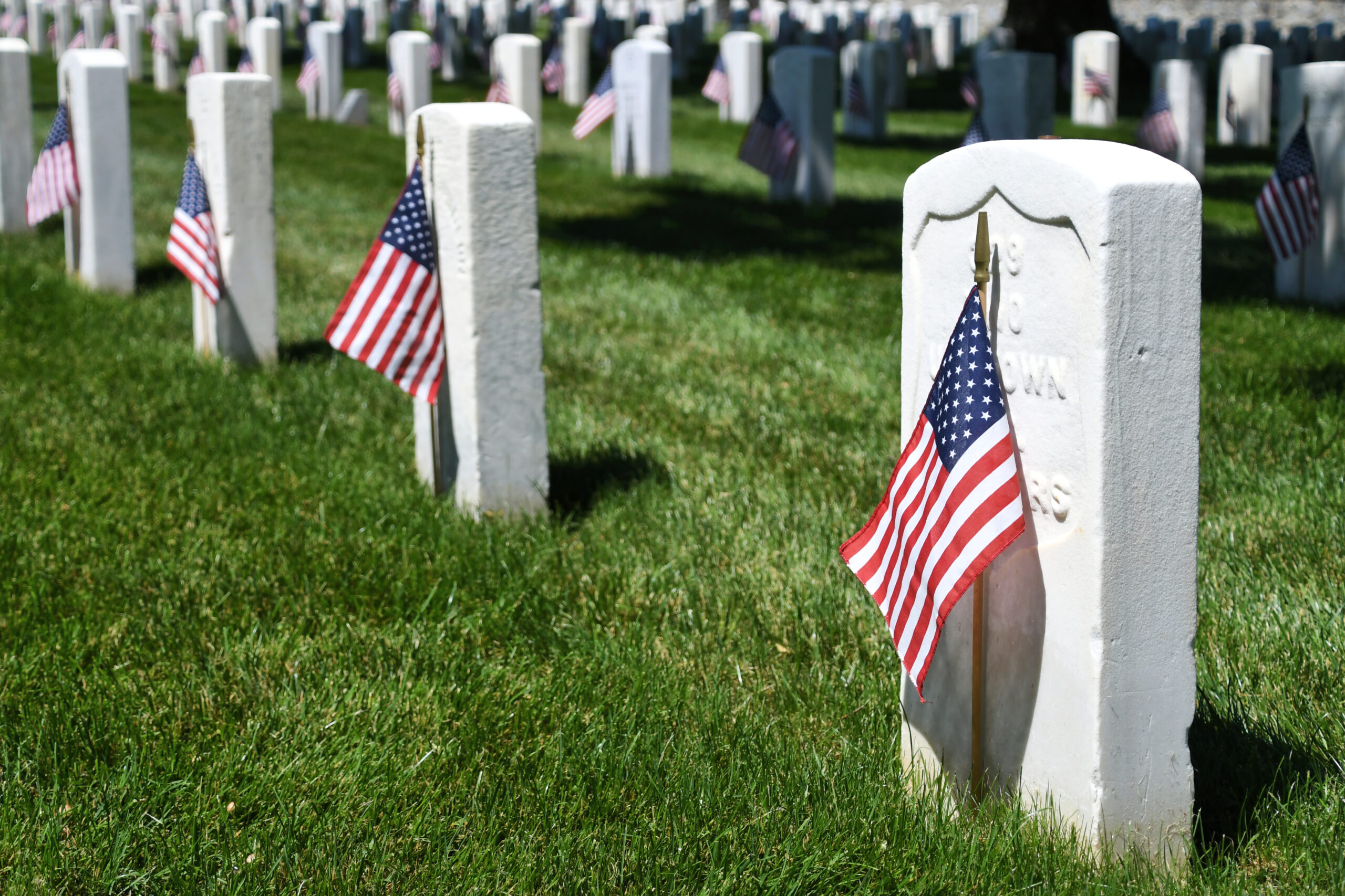Rows of white military gravestones in a grassy cemetery, each marked with a small American flag.