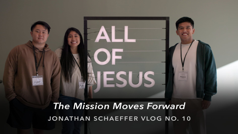 Three people wearing lanyards stand beside a lit sign reading “ALL OF JESUS,” with overlay text: “The Mission Moves Forward” and “JONATHAN SCHAEFFER VLOG NO. 10.”