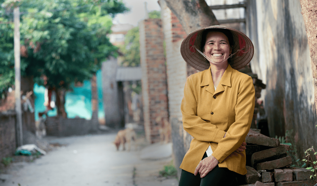 Woman in a yellow jacket and black pants leaning against a stack of bricks in a narrow alleyway with a tree and brick walls