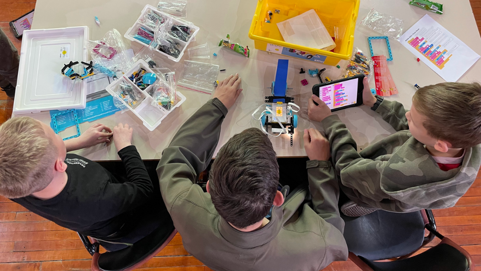 Three kids work on a robotics project at a table with parts, trays, and a tablet showing block coding.