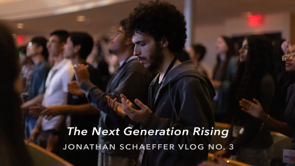 A man with curly hair and a beard stands with open hands in a dimly lit room, surrounded by others in similar reflective postures. Text reads: 'The Next Generation Rising' and 'Jonathan Schaeffer Vlog No. 3'.