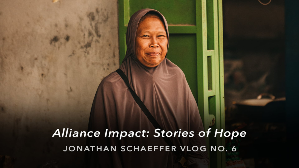 Woman in a long hooded garment standing near a green doorway with text overlay that reads “Alliance Impact: Stories of Hope – Jonathan Schaeffer Vlog No. 6.”