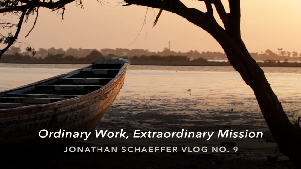 A wooden boat rests at the edge of a calm river at sunset, framed by a silhouetted tree branch. Soft golden light reflects on the water, with overlaid text reading “Ordinary Work, Extraordinary Mission — Jonathan Schaeffer Vlog No. 9.”