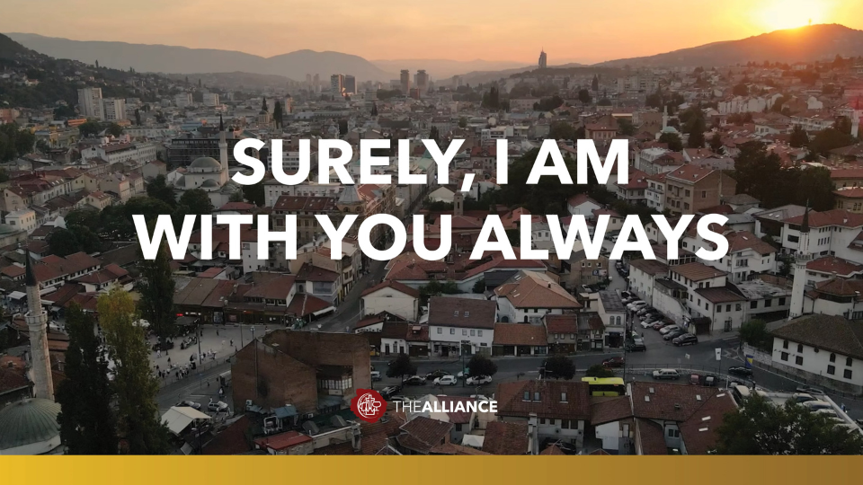 Aerial view of a dense hillside city with red-roofed buildings at sunset, mountains in the background, and warm golden light across the skyline. Large white text in the center reads, “SURELY, I AM WITH YOU ALWAYS,” with a small “THE ALLIANCE” logo at the bottom.
