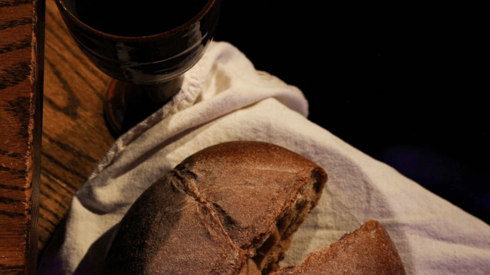 A rustic wooden table with a dark ceramic cup filled with red wine and a round loaf of brown bread partially broken, resting on a white cloth.