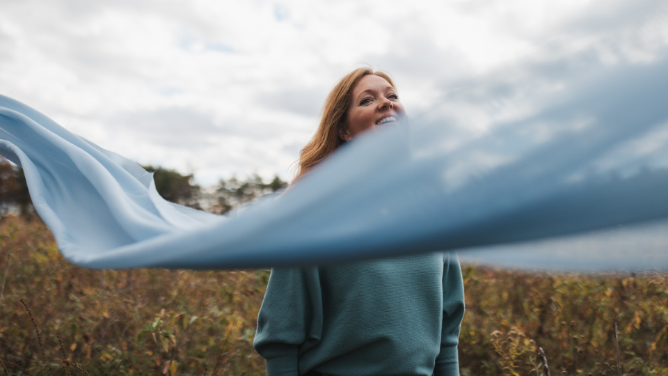 woman standing outdoors in a grassy field wearing a teal sweater, with a light blue fabric flowing across the foreground. The sky is cloudy, and trees are visible in the background.
